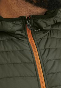 Veste verte matelassée avec une fermeture éclair orange, dotée d'un col haut et d'un tissu texturé, gros plan sur la zone de la fermeture éclair.