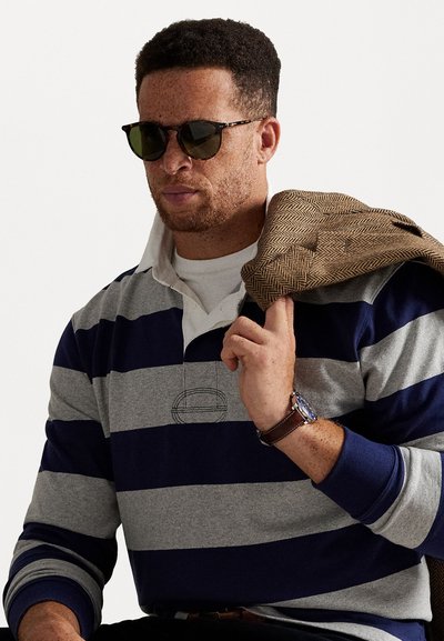 Gestreepte rugbyshirt in grijs en marineblauw, met een witte kraag; houdt een bruin houndstoothjacket over de schouder; draagt donkere zonnebril.