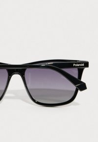 Μαύρα γυαλιά ηλίου Polaroid με σχήμα γάτας, διαθέτοντας φακούς με κλίση από σκούρο μοβ σε ανοιχτό. Λεπτοί σκελετοί με γωνιώδεις κροτάφους.