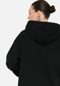 Une femme de dos porte un sweat à capuche noir, affichant une coiffure élégante et des boucles d'oreilles scintillantes. Fond minimaliste.