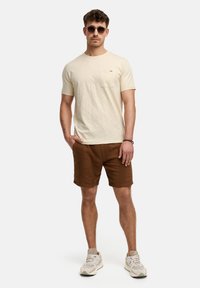 Camiseta de algodón beige con un bolsillo en el pecho, combinada con pantalones cortos marrones texturizados y zapatillas ligeras. Los accesorios incluyen gafas de sol redondas oscuras.