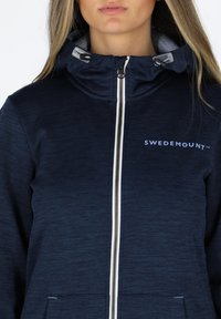 Giacca con zip e cappuccio blu navy in tessuto testurizzato, con zip bianca e logo "SWEDEMOUNT" sul lato sinistro del petto. Presenta tasche laterali.