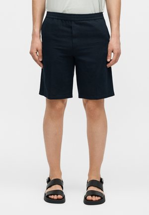 Άνδρας που φοράει μαύρα ελαστικά σορτς στη μέση και μαύρα σανδάλια με διπλό λουράκι στέκεται μπροστά σε ένα μονόχρωμο φόντο.