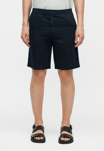 Homme portant un short noir à taille élastique et des sandales noires à double bride, debout devant un fond uni.