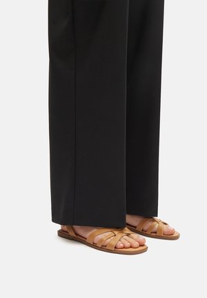 Sandalias de cuero marrón con múltiples tiras superpuestas, suela plana y punta abierta, combinadas con pantalones de pierna ancha negros.