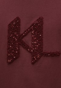 Bordo audums ar izteiksmīgu teksturētu 'KL' logo, kas izveidots no fliteriem, piešķirot mirdzuma un dziļuma variācijas pret gludo fonu.