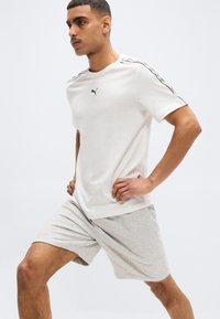 Νέος άνδρας με λευκό T-shirt Puma και γκρι σορτς τεντώνει τα πόδια του, με τα χέρια στη μέση, μπροστά σε απλό ανοιχτό φόντο.