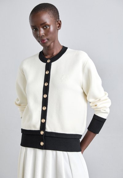 Cardigan en polaire blanche avec des finitions noires, boutons dorés sur le devant et manches côtelées. Détail du logo en relief sur la partie gauche de la poitrine.