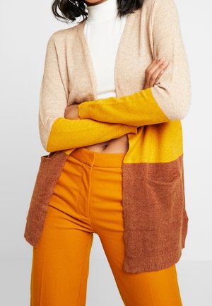 Strickjacke in Beige mit gelbem und braunem Colorblocking, mit gerippten Bündchen und Taschen. Kombiniert mit orangefarbenen High-Waist-Hosen.