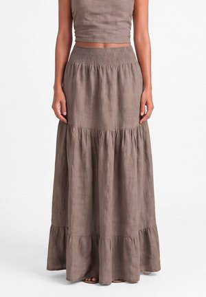 Kobieta w brązowej, wielowarstwowej maxi spódnicy z marszczonym pasem i dopasowanym krótkim topem bez rękawów na białym tle.