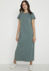 Green cotton maxi dress with short rolled sleeves, straight cut, side slits, and soft texture, paired with white athletic sneakers.