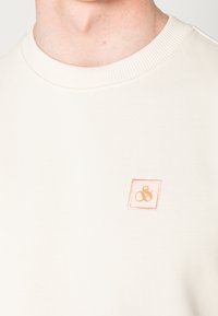 Sudadera crema con cuello de canalé, que presenta un pequeño logotipo cuadrado de durazno bordado en el pecho. Textura suave, diseño casual.