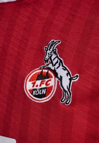 Rødt stoff har et brodert logo av 1. FC Köln, som skildrer en hvit geit med svarte detaljer, omkranset av en sirkulær rød og hvit kant.
