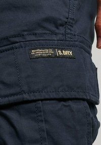Pantaloni cargo in tessuto blu navy con finitura testurizzata. Presentano un'etichetta dorata e nera con la scritta "S.DRY" e ulteriori dettagli del marchio.