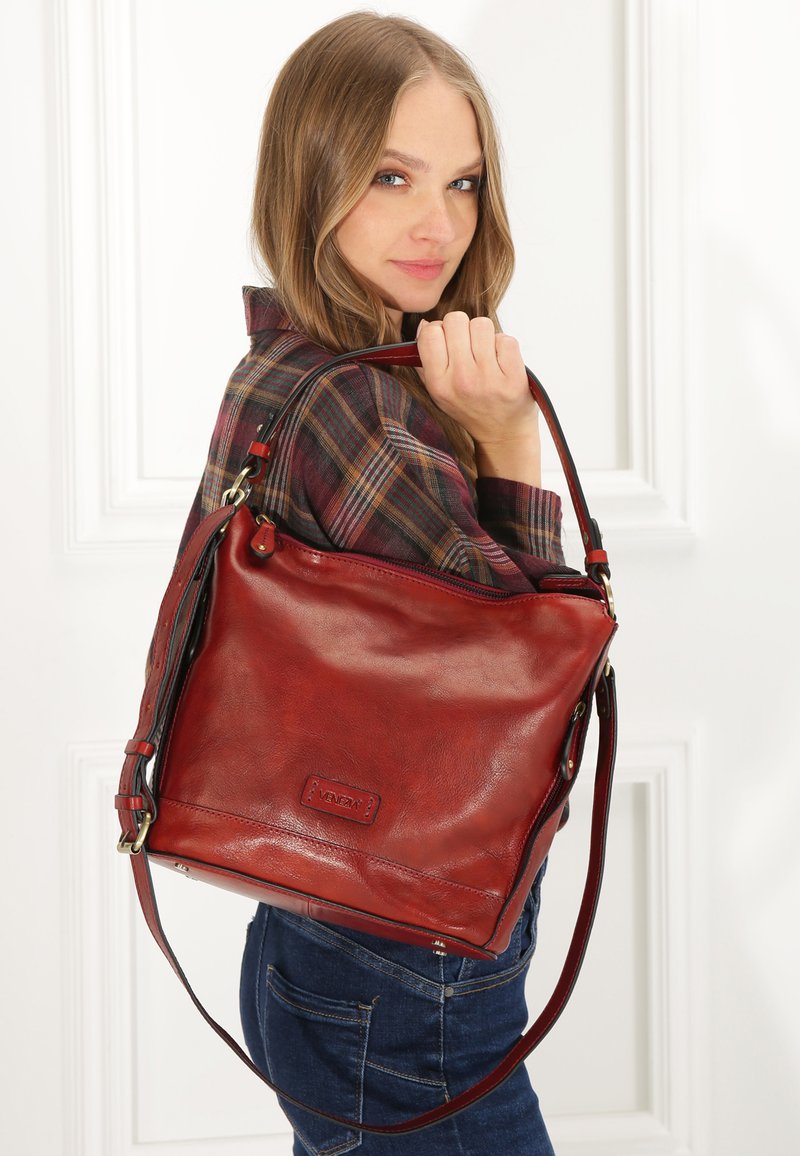 Sac à main en cuir rouge avec un design décontracté, doté d'une seule bandoulière, d'une fermeture éclair et d'une petite étiquette de marque à l'avant.