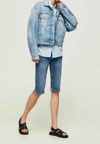 Veste en jean bleu clair, avec deux poches avant et une coupe courte. Associée à une chemise bleu clair et un short en jean arrivant aux genoux. Sandales noires.