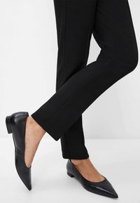 Chaussures plates en cuir noir avec un bout pointu et un petit talon, portées avec un pantalon noir ajusté, mettant en avant un design épuré et minimaliste.