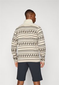 Pullover beige à motifs avec un col haut, mettant en avant des designs géométriques noirs et marron, associé à un short sombre. Tissu texturé.