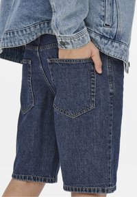 Tummansiniset farkkushortsit, joissa on rento istuvuus, useita taskuja ja hieman risat helmat. Yhdistettynä vaaleansiniseen farkkutakkiin.