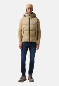 Beige polstret vest med høj krave, lynlåslommer og logo-patch, parret med en beige strikhue, beige sweater, blå jeans og brune sneakers.