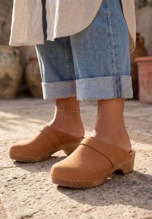 Pieds portant des sabots en cuir beige avec talons en bois, associés à un jean bleu retroussé et de fines chevillères en or sur un sol en pierre en extérieur.