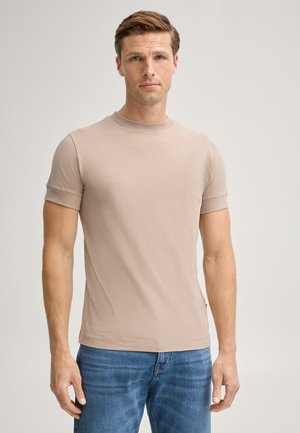 Hombre con una camiseta lisa de manga corta beige y vaqueros azules, de pie frente a un fondo liso y claro, mirando hacia adelante.