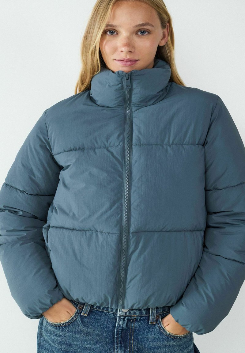 Kurtka puchowa w kolorze niebiesko-zielonym, z wysokim kołnierzem, zamkiem błyskawicznym i pikowanym wzorem. Wykonana z lekkiej, teksturowanej tkaniny.