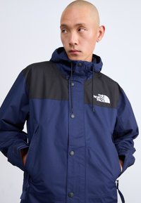 Tamsiai mėlyna vandeniui atspari striukė su juoda viršutine dalimi, priekiniais mygtukais, gaubtu ir šoninėmis kišenėmis. Ant krūtinės yra logotipas, o rankogaliai elastiniai.