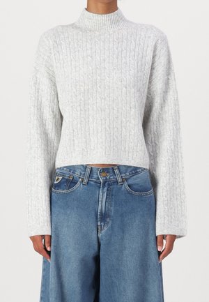 Personne portant un pull à col roulé en maille torsadée gris clair et un jean bleu ample, debout devant un fond uni.