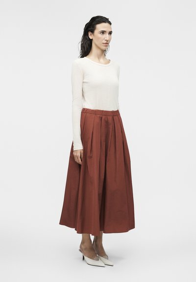 Γυναίκα που στέκεται φορώντας λευκό μακρυμάνικο τοπ, πλισέ midi φούστα σε σκουριά απόχρωση με ψηλή μέση και λευκά παπούτσια με μυτερή μύτη.