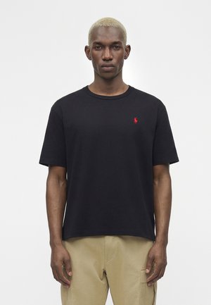 Άνδρας με κοντά ξανθά μαλλιά, φοράει απλό μαύρο T-shirt με ένα μικρό κόκκινο λογότυπο στο στήθος και μπεζ παντελόνι, στέκεται μπροστά σε λευκό φόντο.