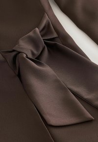 Teren din fibră de culoare maro cu o textură netedă, având un detaliu evidențiat sub formă de fundiță la nivelul taliei. Designul este elegant și bine structurat.