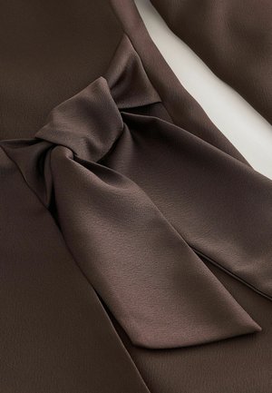 Hnědá tkanina s hladkou texturou, která k sobě přitahuje pozornost díky výraznému detailu mašle v pase. Design je elegantní a strukturovaný.