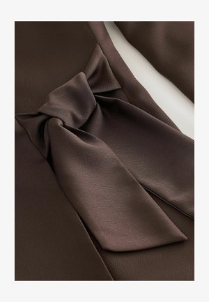 Hnědá tkanina s hladkou texturou, která k sobě přitahuje pozornost díky výraznému detailu mašle v pase. Design je elegantní a strukturovaný.