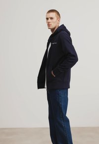 Felpa con cappuccio blu navy e zip frontale, tasche laterali e dettaglio del logo bianco. Indossata con jeans blu a gamba dritta. Tessuto liscio.