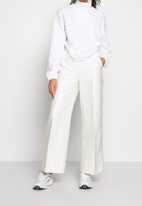 En person klädd i en vit zip-up sweatshirt, vida vita byxor och vita sneakers, står mot en enkel vit bakgrund.