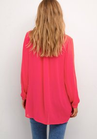 Blusa a maniche lunghe di un vivace colore rosa, realizzata in tessuto leggero con una vestibilità ampia e orlo curvato, abbinata a jeans blu aderenti.