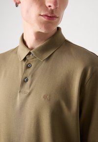 Polo shirt vert olive avec une patte de boutonnage à deux boutons, manches courtes et logo discret. Tissu doux, texture lisse et design de col classique.