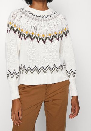 Personne portant un pull en maille blanc avec un motif zigzag multicolore sur la poitrine et les manches, associé à un pantalon marron.