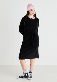 Black knitted dress with long sleeves, tie waist detail, and knee-length cut, paired with black Converse sneakers and a pink beanie.