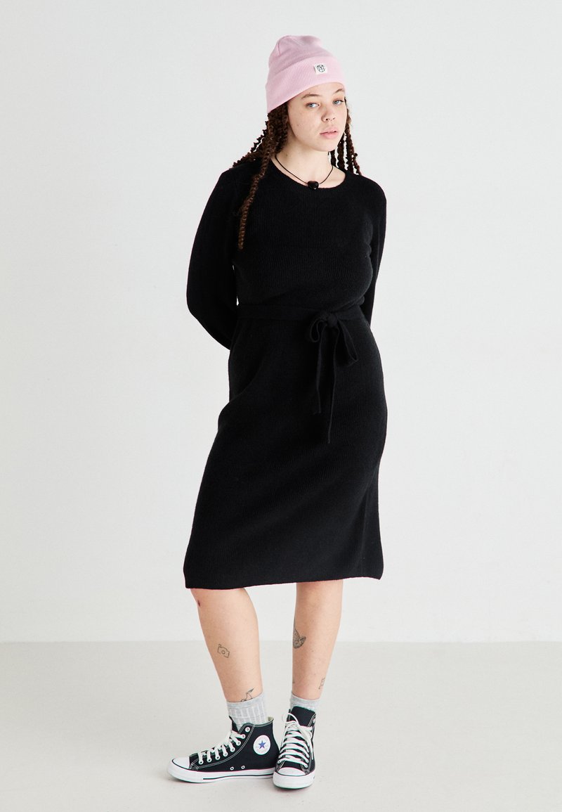 Czarna dzianinowa sukienka z długimi rękawami, z detalem przewiązania w talii i długością do kolan, połączona z czarnymi trampkami Converse i różową czapką beanie.