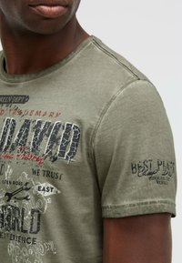 Olive green T-shirt in soft cotton with a crew neck, featuring graphic prints in black and red, including text and icons on the front and sleeve.