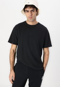 Tricou negru din bumbac, cu o croială relaxată, având un logo debossat pe piept. Asortat cu pantaloni jogger negri și o pălărie tip bucket de culoare deschisă.