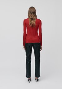 Femme avec de longs cheveux ondulés portant un pull rouge côtelé, un pantalon en cuir noir et des chaussures à talons noirs, se tenant de dos sur un fond gris.