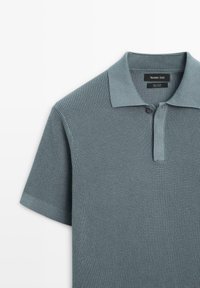 Polo en coton vert texturé à manches courtes, avec un col plié et deux boutons au niveau du cou. Étiquette de la marque visible à l'intérieur du col.