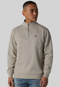 Beige hettejakke med glidelås og høy krage, med horisontale sømdetaljer, laget av mykt materiale. Den har en liten logo på brystet.