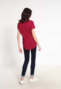 Kvind med skulderlangt mørkt hår iført en rød kortærmet top, sorte skinny jeans og hvide slip-on sko, stående med ryggen til.