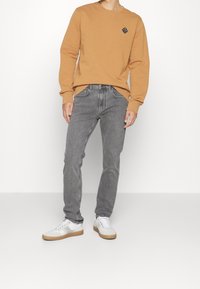 Hnědá mikina s malým černým logem, spárovaná se šedými džíny a bílými teniskami s gumovými podrážkami. Jednoduché, ležérní oblečení.