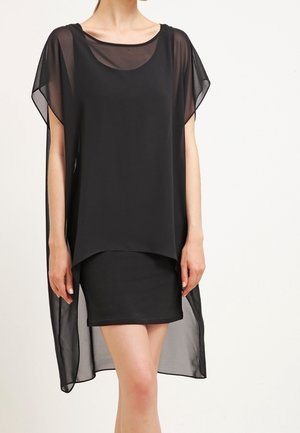 Frau trägt ein schwarzes, ärmelloses Kleid mit einer transparenten, lockeren Außenschicht mit kurzen Ärmeln und einem vorn kürzeren, hinten längeren Saum.