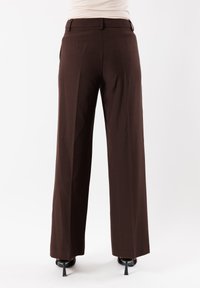 Pantaloni marroni a gamba larga con una texture liscia, caratterizzati da pieghe anteriori e una vita standard. Tacchi neri visibili sotto l'orlo.